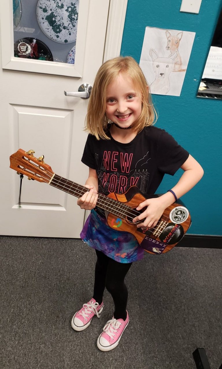 A young girl stands in a music school against a green wall holding a ukulele and smiling at the camera