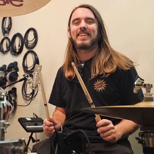 Man with long light brown hair sits at a drum set in a professional recording studio, holding drumsticks