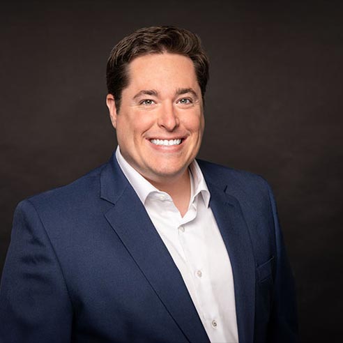 portrait of a smiling man with close-cut brown hair, wearing a casual business suit against a grey portrait studio backdrop