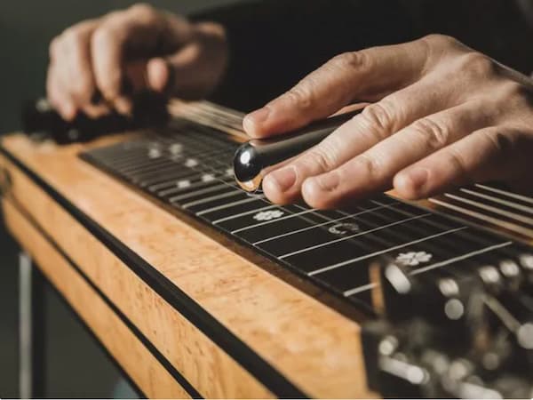 closeup of two hands on a pedal steel guitar. One hand is resting near the strings, while the other is holding a steel slide