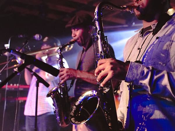 Three brass players performing on stage. Two are playing saxophones, one is playing a trombone