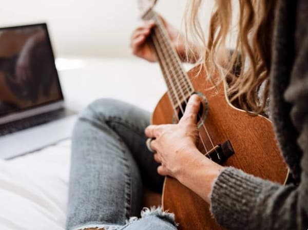 Person with long hair sitting cross-legged on top of a bad strumming a ukulele, looking at a laptop
