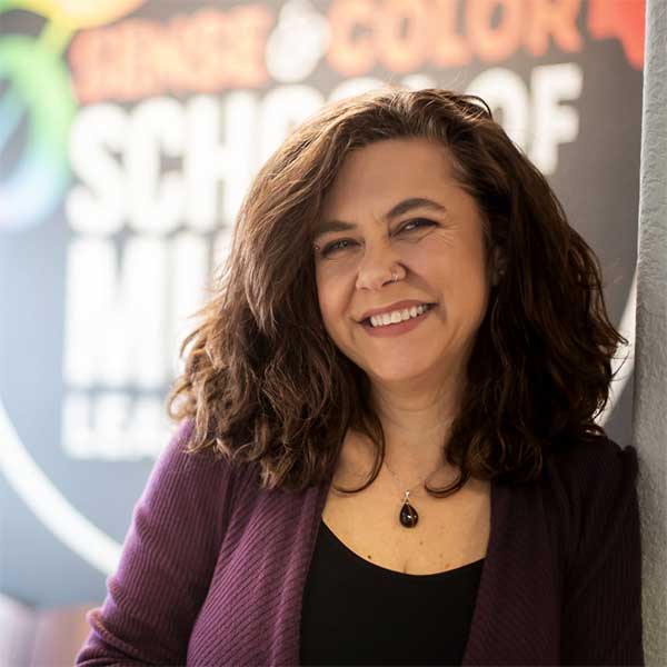 Woman with dark curly hair smiles, while standing in front of a brightly painted wall with the Sense & Color School of Music logo