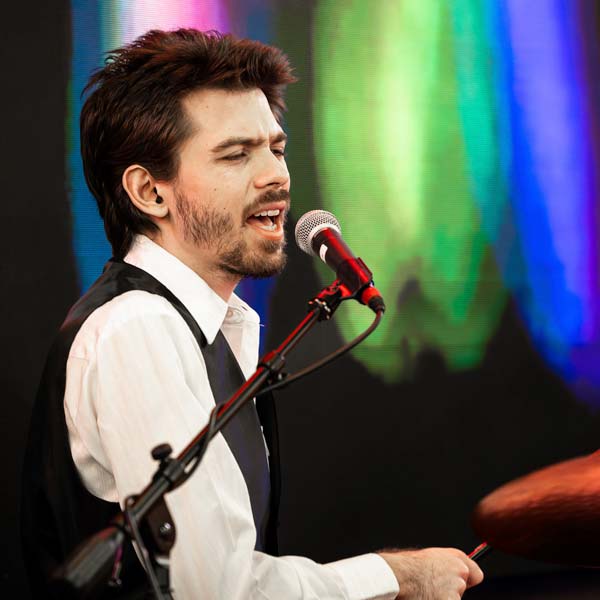 Man in his early twenties with short brown hair and a light beard sings into a mic while playing drums, against a rainbow-colored stage lighting backdrop